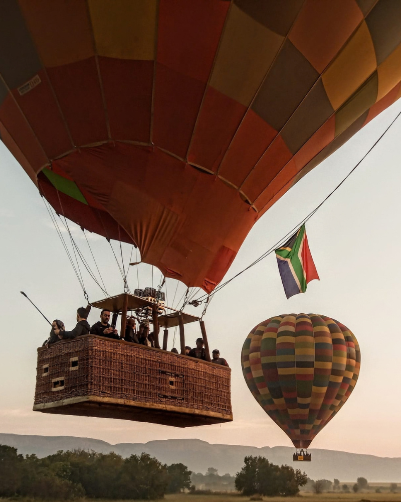 Hot air balloon carrying passengers beneath a South African flag, with a second balloon in the background